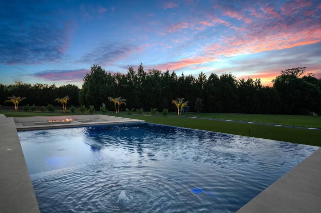 A backyard pool glowing with reflections of the sunset sky, framed by trees and a sleek modern deck.