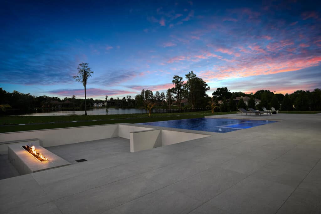 A backyard pool reflecting a vibrant sunset, with surrounding white tiles and lush landscaping completing the scene.