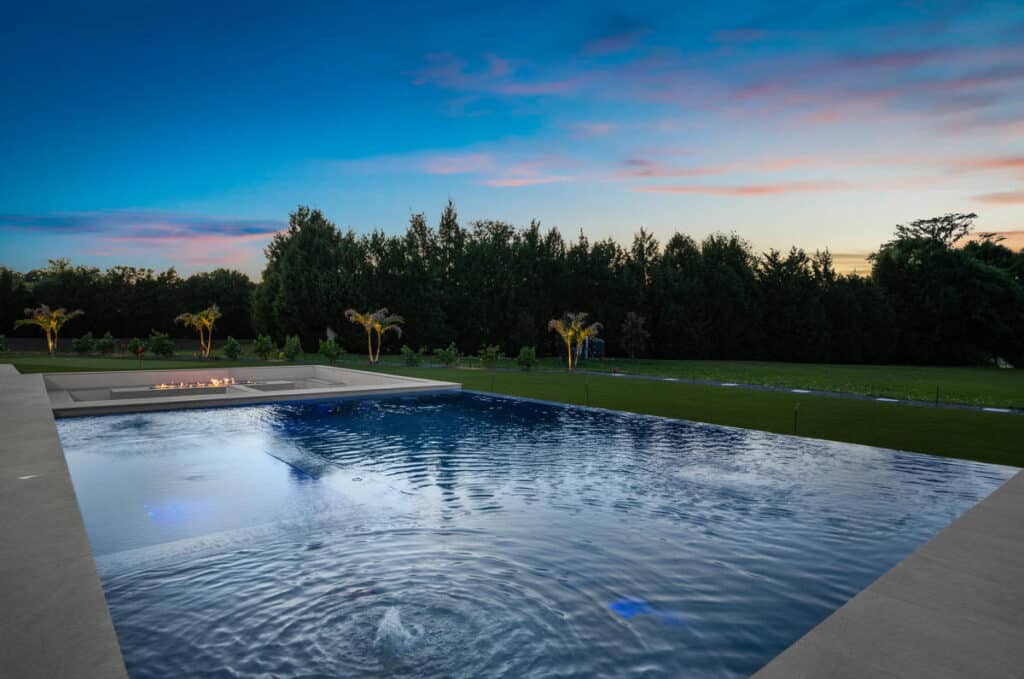 A backyard view of a rectangular pool surrounded by trees and shrubs, with the soft glow of a setting sun painting the sky in pastel colors.
