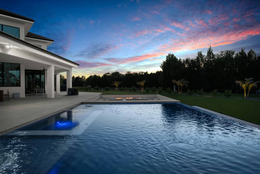 A beautiful pool reflecting a twilight sky with pink and purple hues, surrounded by contemporary architecture and trees.