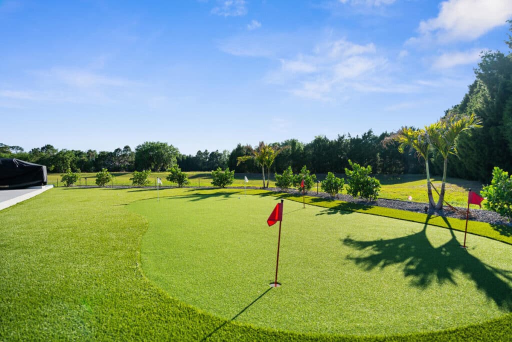A bright green putting green framed by trees, with a red practice flag under a sunny blue sky.