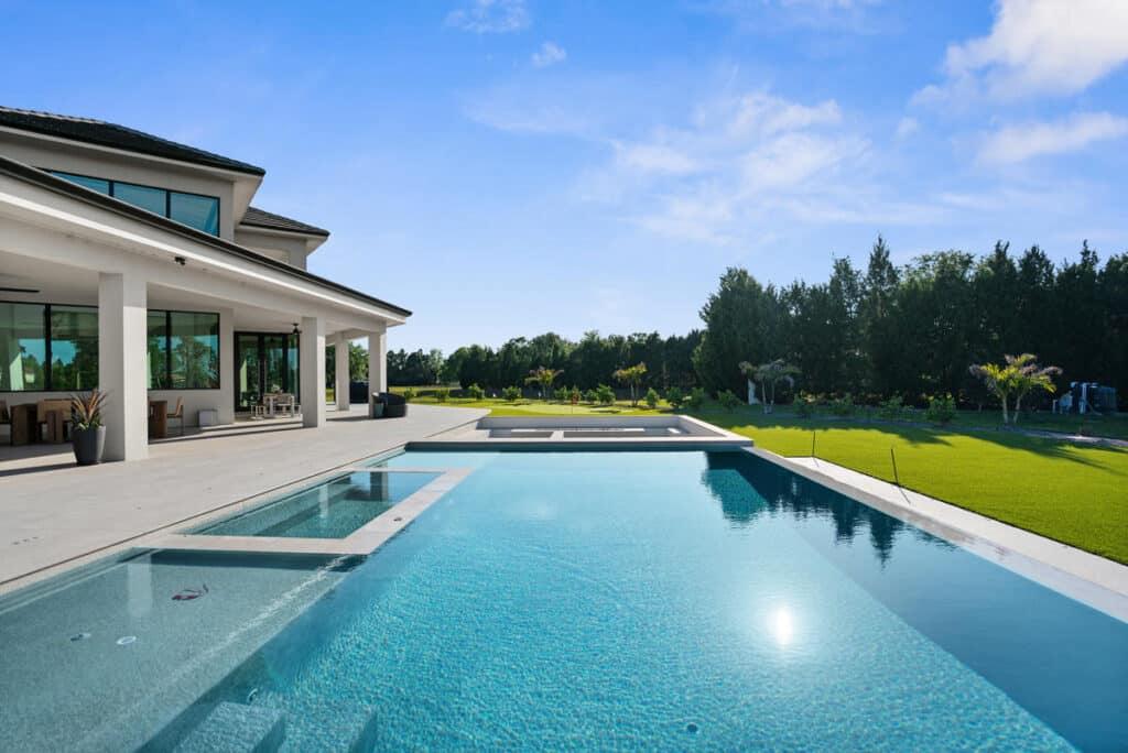 A clear blue swimming pool reflecting the sky, surrounded by a modern house and well-manicured greenery.