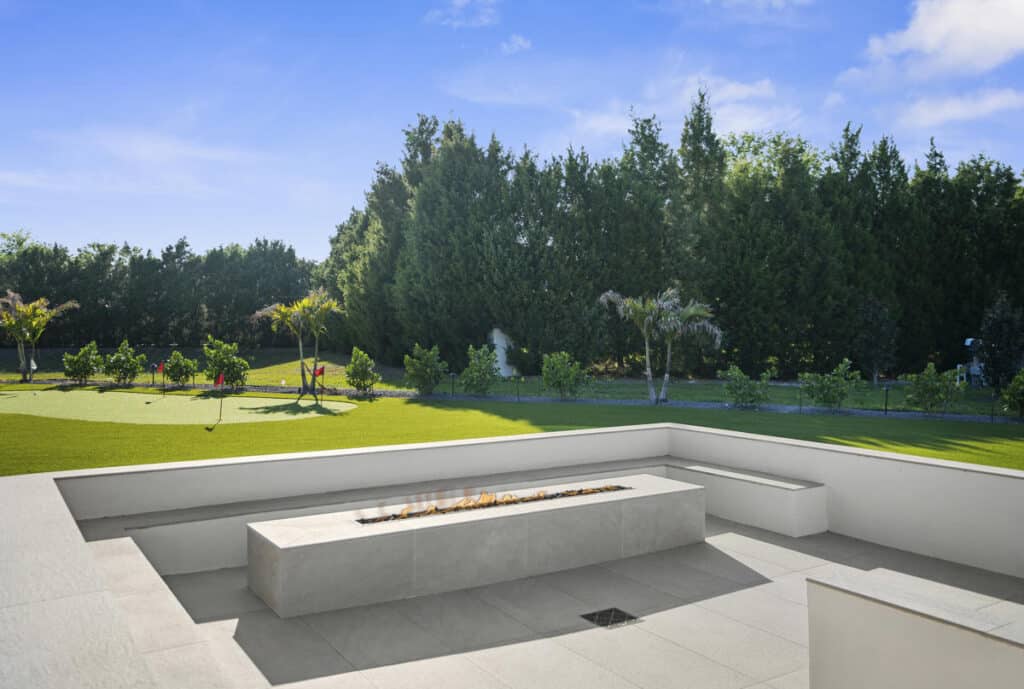 A crisp white concrete bench and fire pit facing a grassy backyard area with tall trees and a peaceful atmosphere.