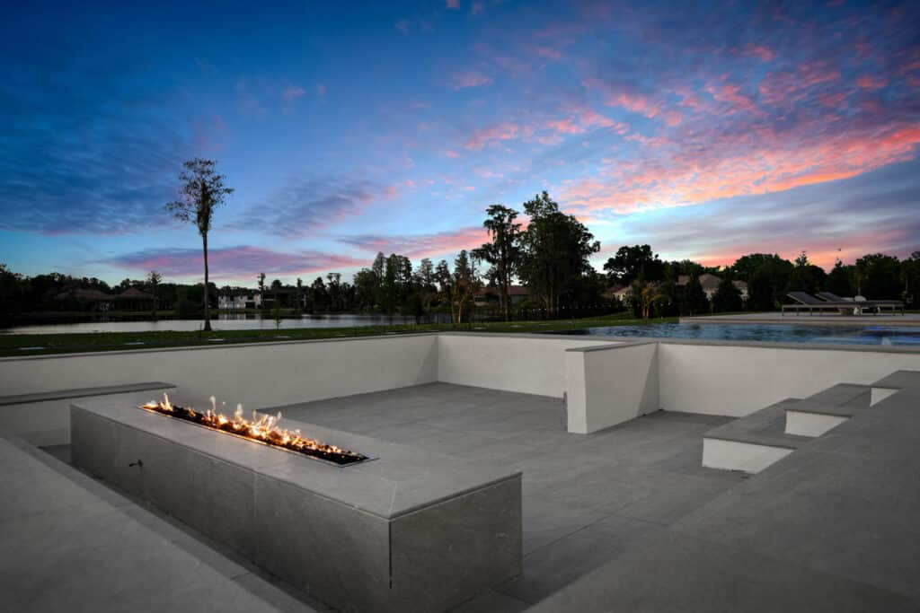 A glowing fire pit surrounded by smooth concrete seating, framed by a modern pool and lush green landscape at sunset.