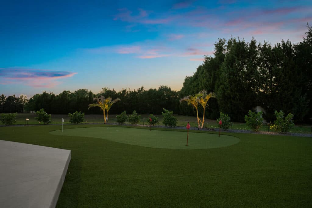 A lush green backyard putting green under a vibrant sunset sky, framed by low shrubs and ambient lighting.