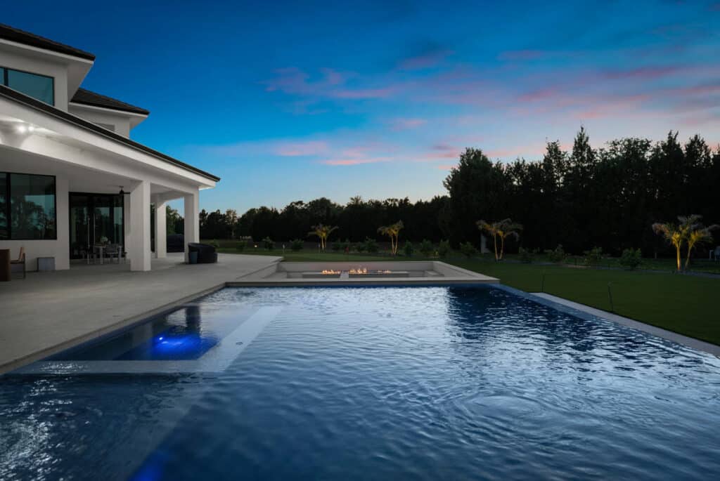 A luxurious modern home with a pool in the foreground, reflecting the warm hues of the sunset sky and surrounded by sleek architecture.