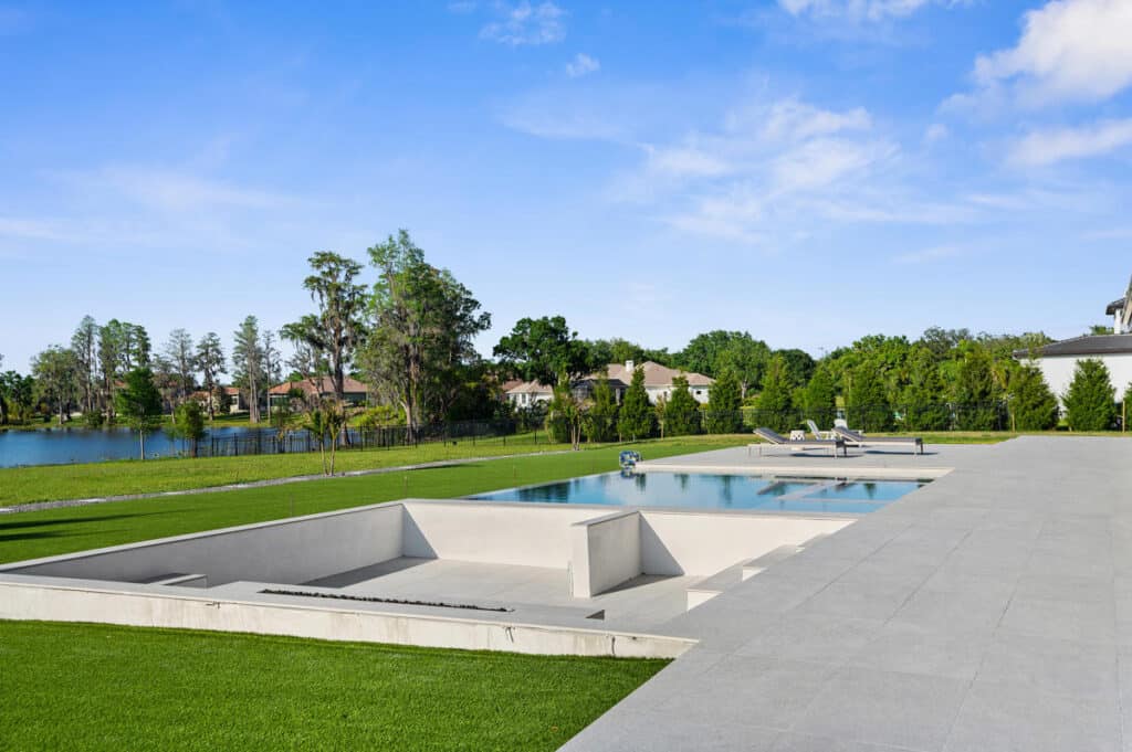 A minimalist pool deck and sunken fire pit with scenic views of a tree-lined property and clear blue skies.
