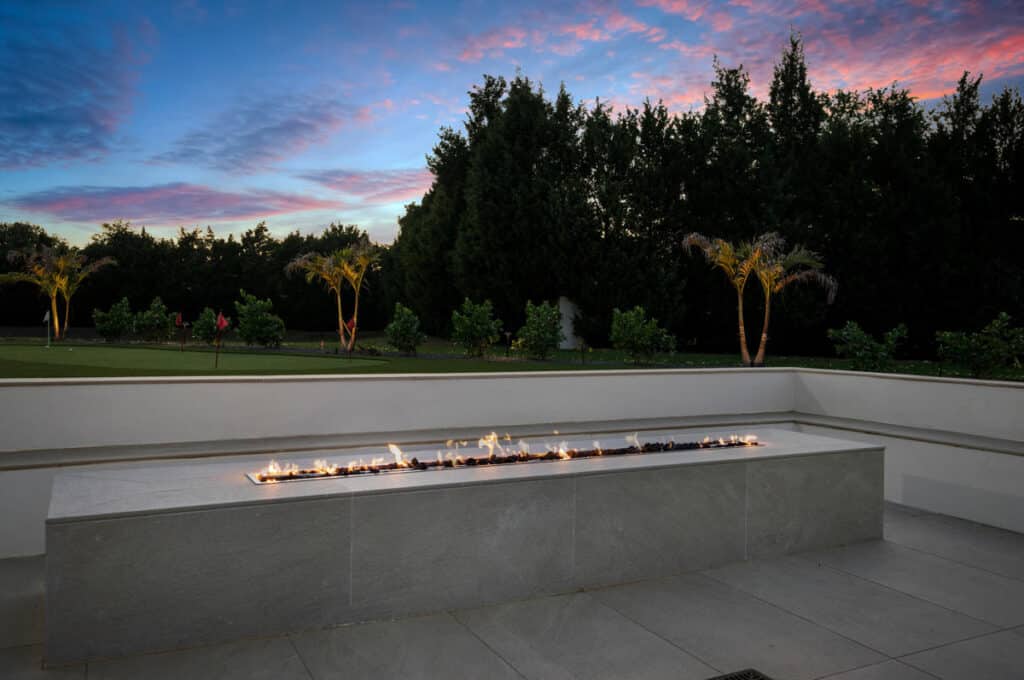 A modern fire feature framed by vibrant green trees and undergrowth, set against the backdrop of a sunset sky.