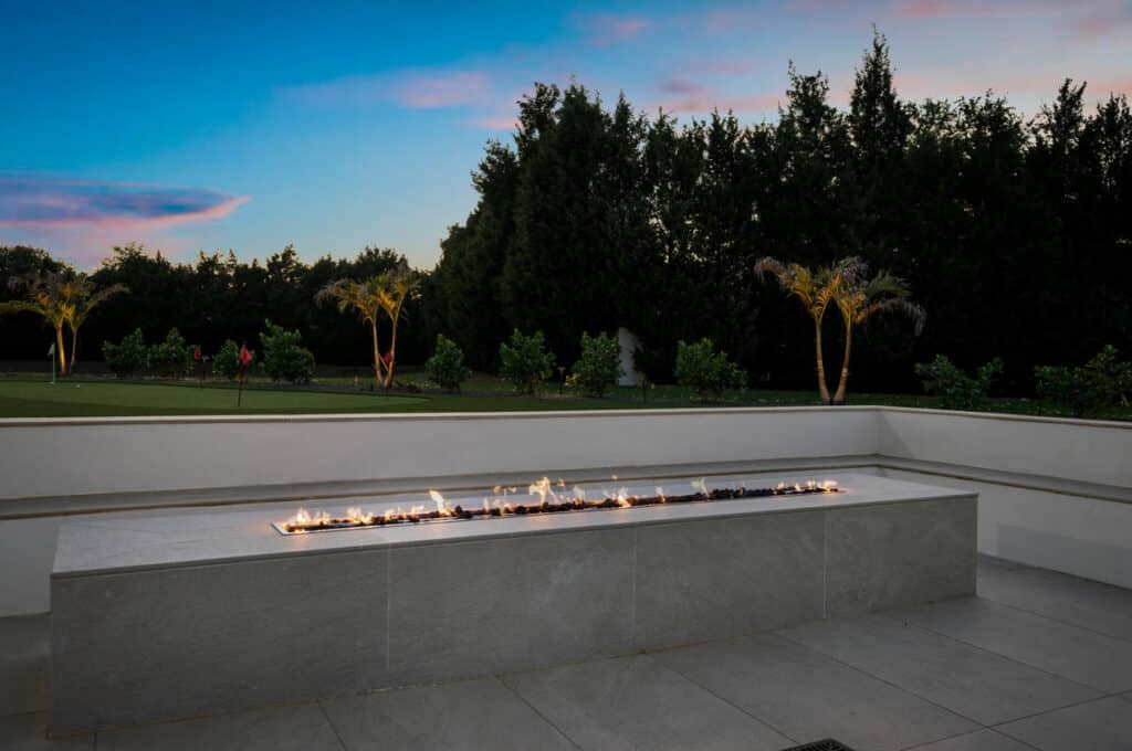 A panoramic view of a modern fire feature surrounded by trees with an expansive sunset sky overhead.