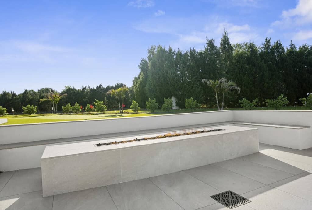 A polished backyard fire feature along a white concrete bench, facing an open lawn with trees in the distance.
