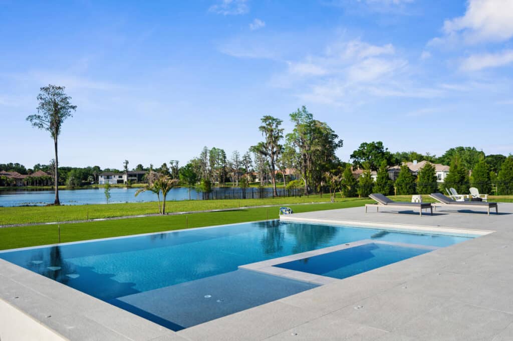 A rectangular pool surrounded by a modern white patio, reflecting the bright blue sky and dotted with greenery.