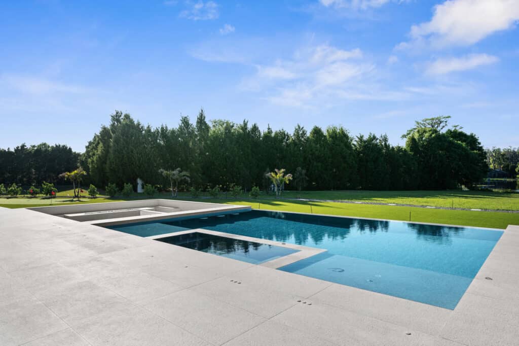 A rectangular pool with crystal-clear water and a modern white patio, surrounded by green trees and a sunny sky.