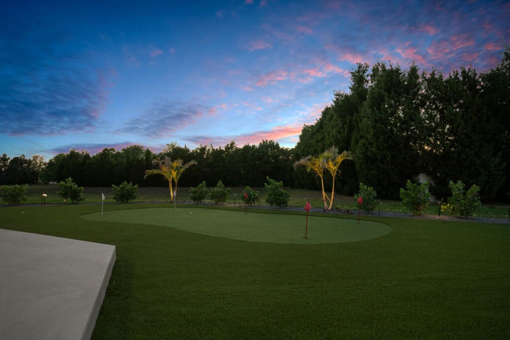 A serene backyard putting green glowing under twilight skies, surrounded by trees and illuminated landscaping features.
