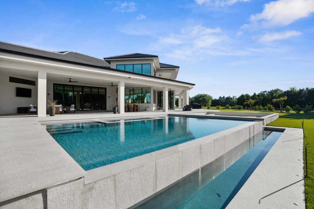 A spacious backyard featuring a rectangular pool and a two-story modern house, surrounded by lush green lawn and trees.
