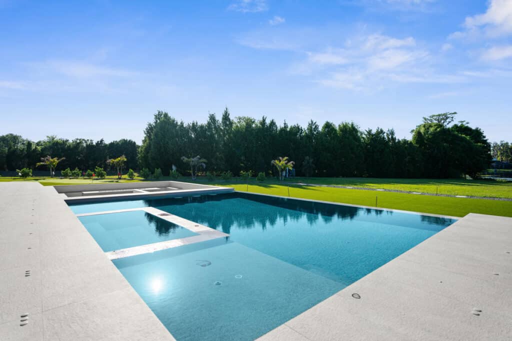 A sunny poolside scene with a clear blue pool, white patio tiles, and a well-maintained green lawn.