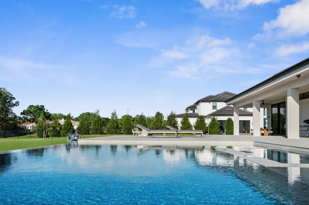 A wide view of a grassy backyard with a swimming pool and a modern home in the background, set against a clear blue sky.