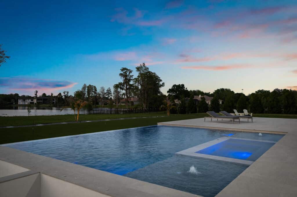 An elegant modern backyard pool reflecting a deep sunset, with tall trees and a minimalist concrete patio framing the scene.