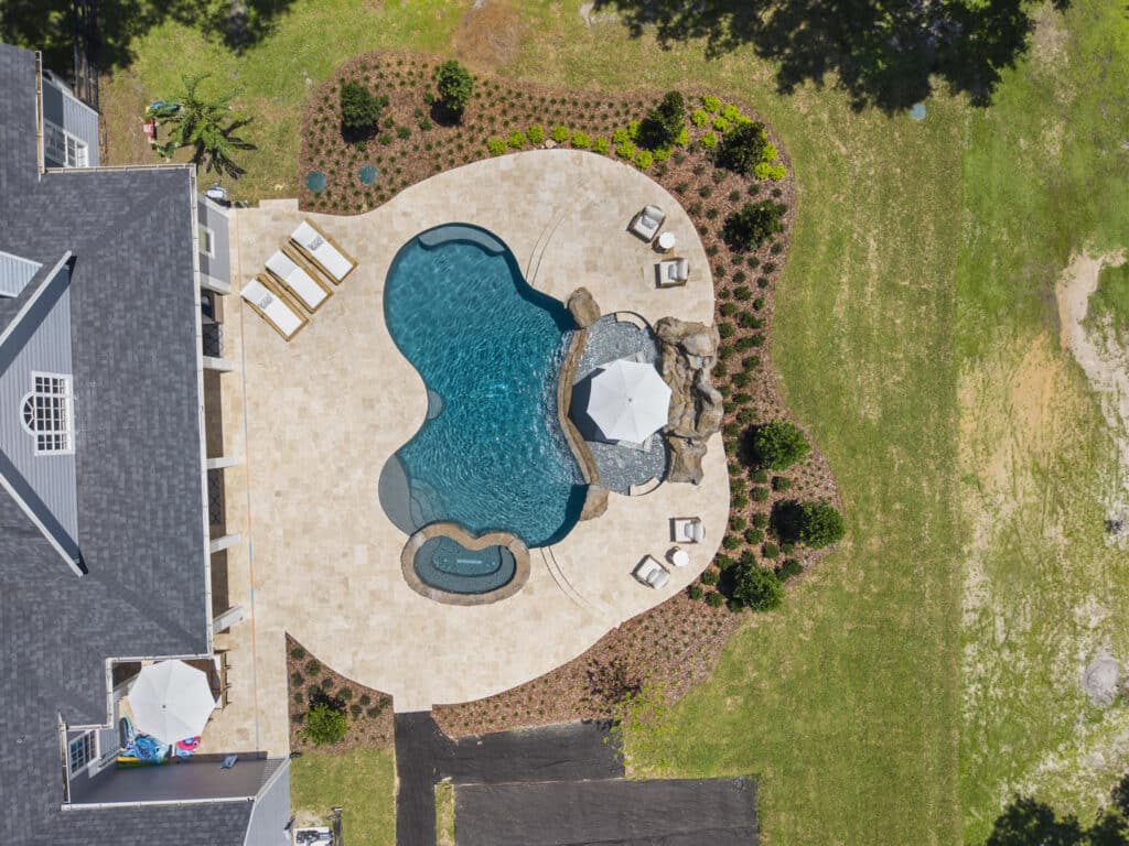 Drone shot of a custom pool with sun shelf, spa, and lounge areas, set in a spacious backyard with landscaped border.
